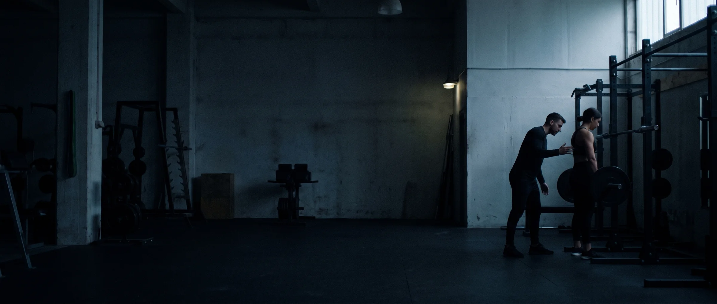 A coach cueing a client beside a squat rack in a dark gym, lit by one cool window